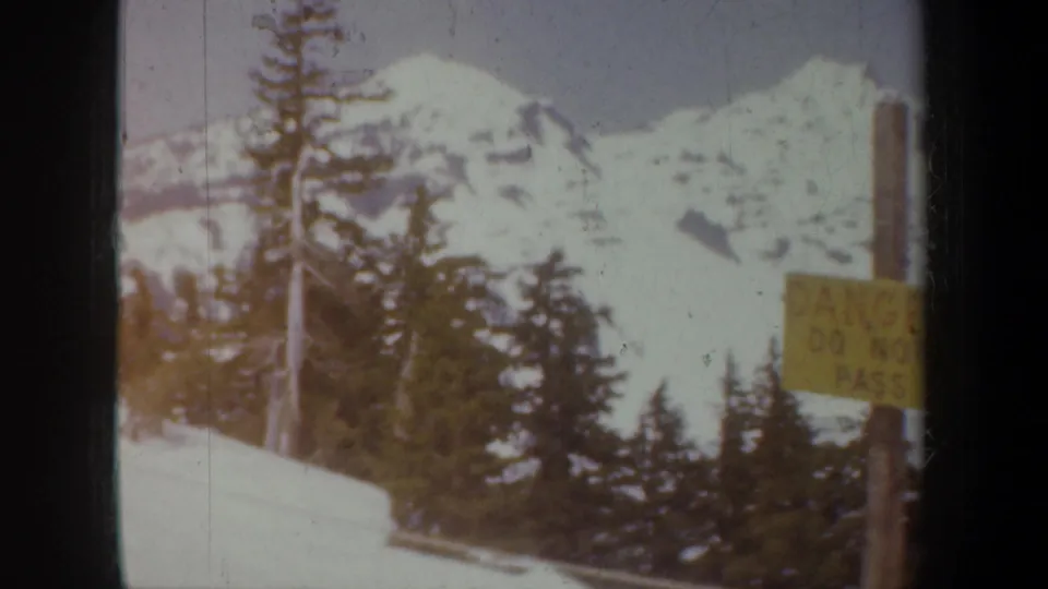 1957 Oregon home-movie frame showing snowy mountains, dark evergreen trees, and a yellow danger warning sign in a winter roadside view.