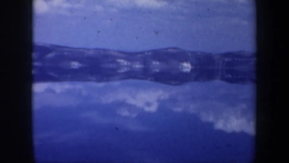Mid-pan frame from a 1957 Oregon home movie showing blue water or ice and a dark horizontal shoreline or mountain ridge.