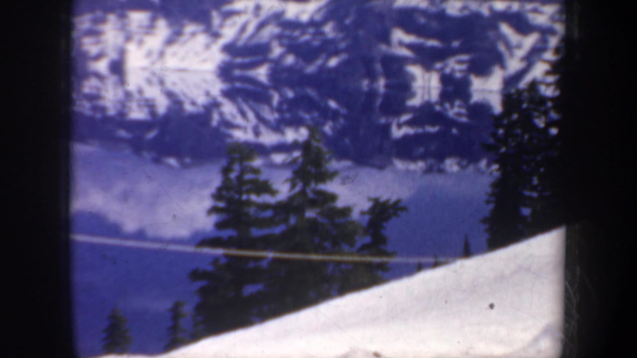 1957 8mm frame from Oregon-area home-movie footage showing snow, evergreens, a blue lake, and distant mountain ridges.