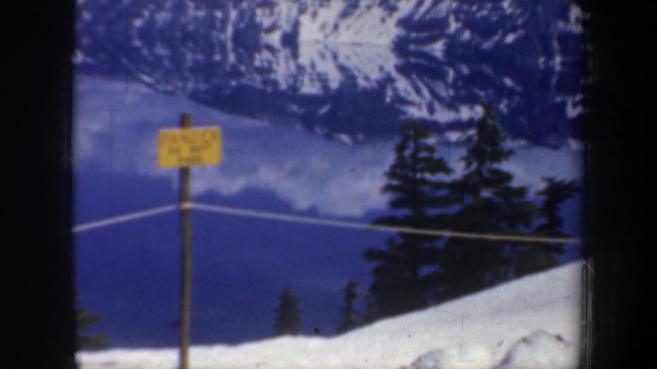 Yellow caution sign beside a cable line, with snow, evergreens, lake, and mountains behind it.