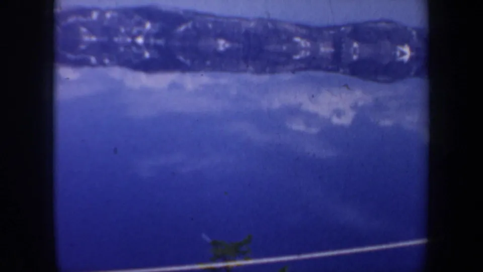 Blue-tinted 1957 8mm frame showing a snowy lakeside area, distant ridge, and a cable line across the lower view.
