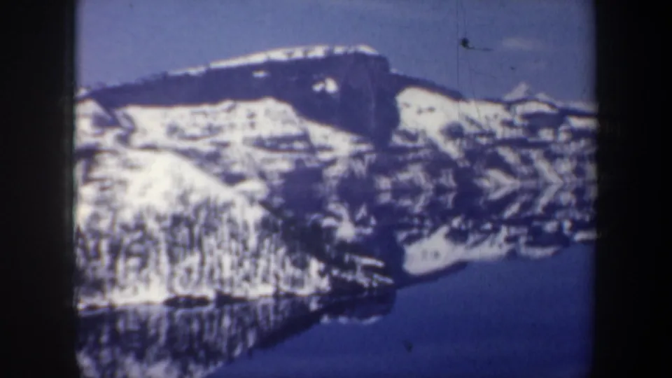 1957 Oregon 8mm frame showing a snow-covered ridge and its reflection in a calm blue lake.