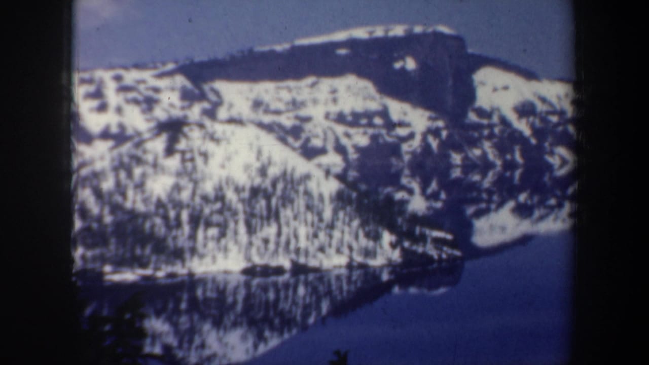 1957 8mm frame of snowy mountain slopes above a calm lake, with dark trees at the lower edge and visible film gate borders.