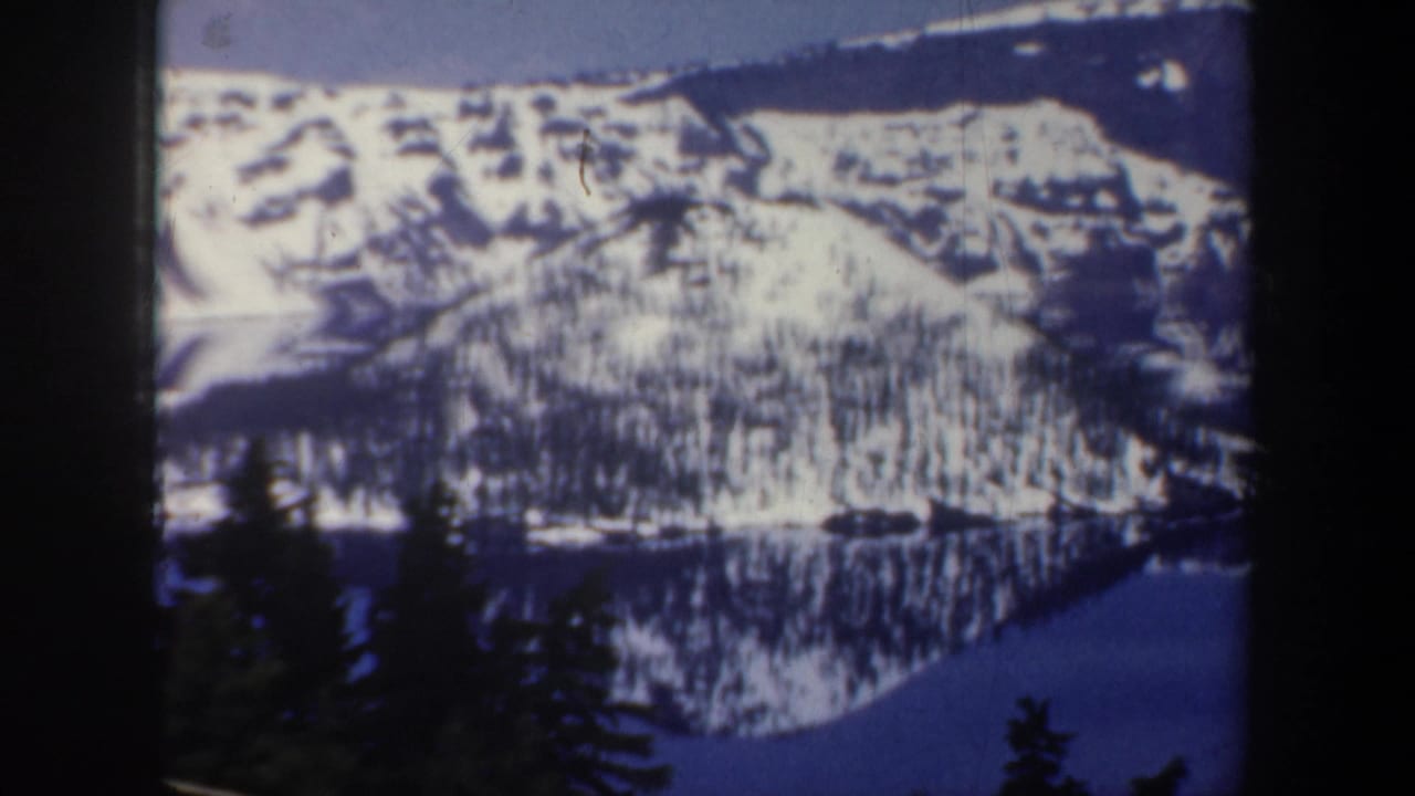 Snowy ridgeline and wooded slope reflected in a calm lake.