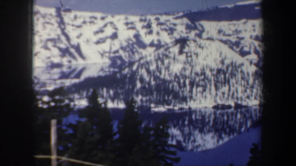 1957 8mm frame of a snow-covered hillside reflected in blue lake water, partly framed by dark evergreen trees and film edges.