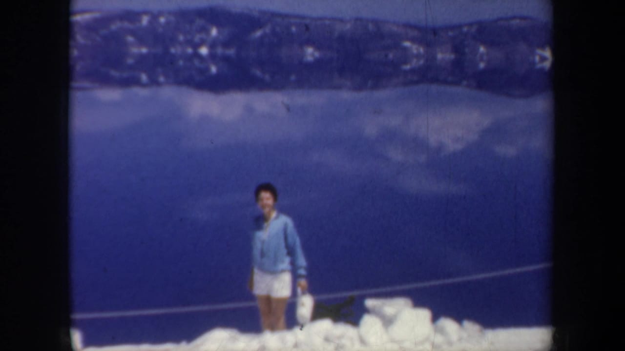 Man in a blue top and white shorts standing on snow above a blue lake or valley.