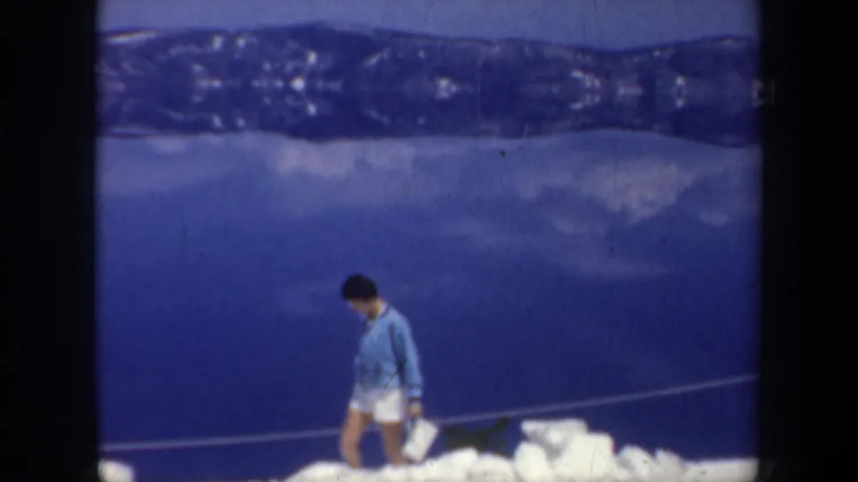 1957 Oregon home-movie frame showing an adult man on a snowy edge, head lowered, with a blue lake or valley backdrop and distant ridge.