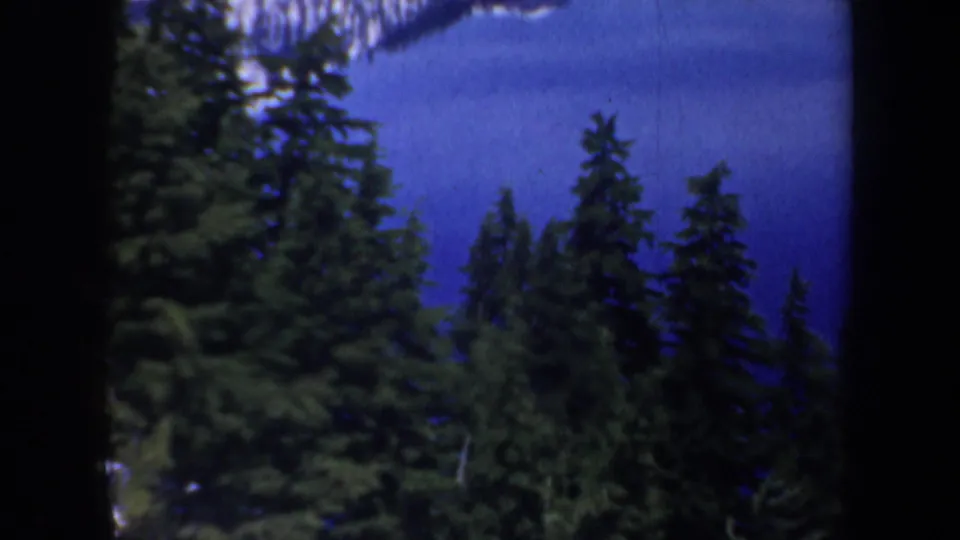 1957 Oregon 8mm frame with evergreen trees in the foreground and a calm blue lake behind them.