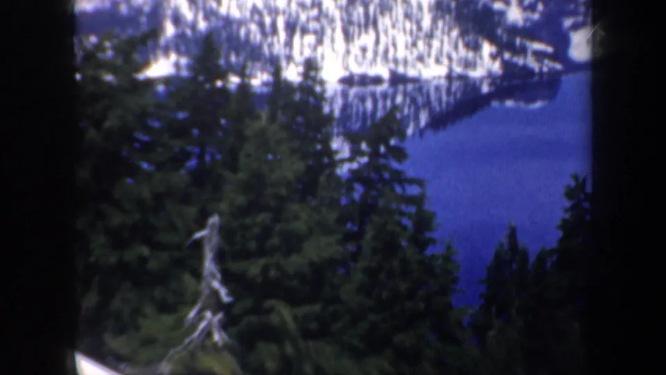 1957 Oregon 8mm view of conifers overlooking a blue lake with snowy mountain terrain beyond.