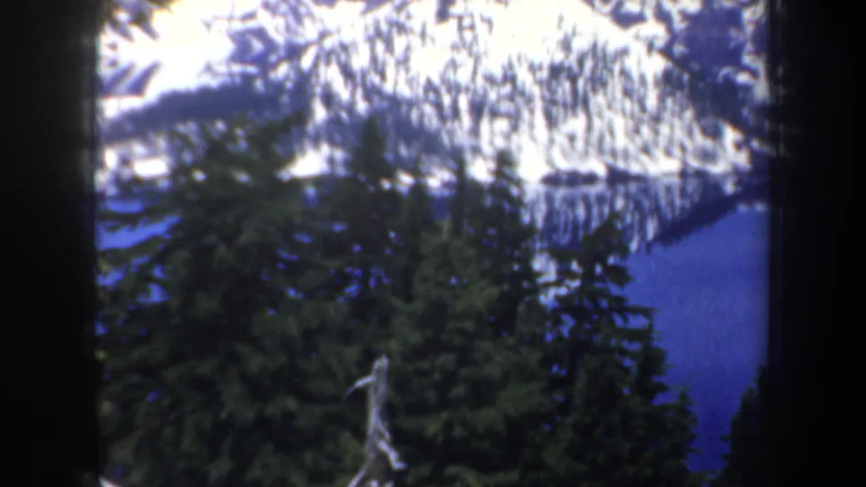 1957 Oregon 8mm frame showing a snow-covered mountain and blue lake behind dark evergreen trees.