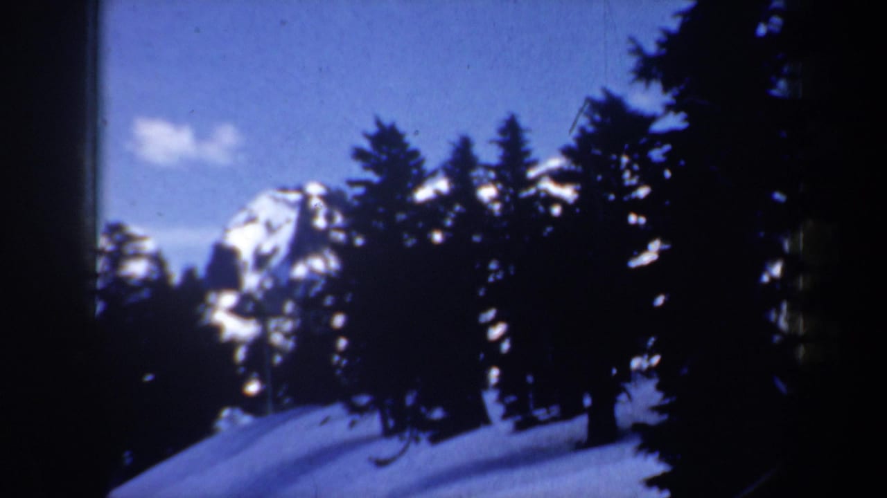 1957 Oregon 8mm frame with dark evergreens, a snow-covered slope, and a distant mountain peak under blue sky.
