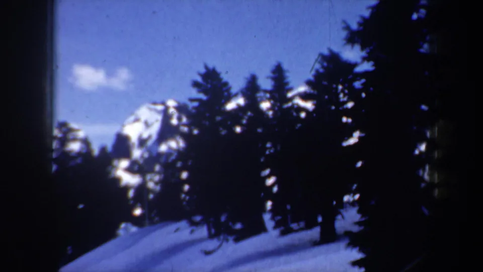 1957 Oregon 8mm frame with dark evergreens, a snow-covered slope, and a distant mountain peak under blue sky.