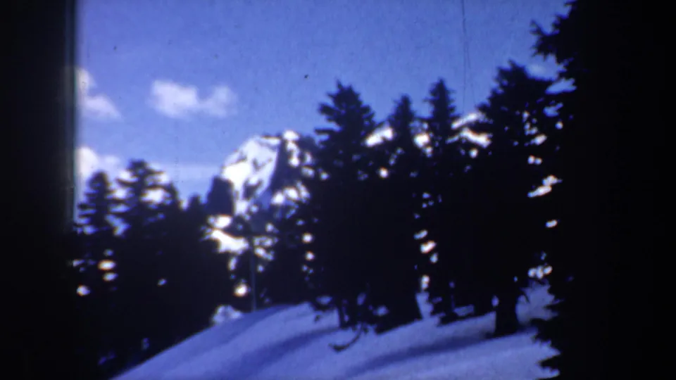 1957 Oregon 8mm frame showing evergreen trees crossing a snowy hillside with distant mountain peaks in the background.