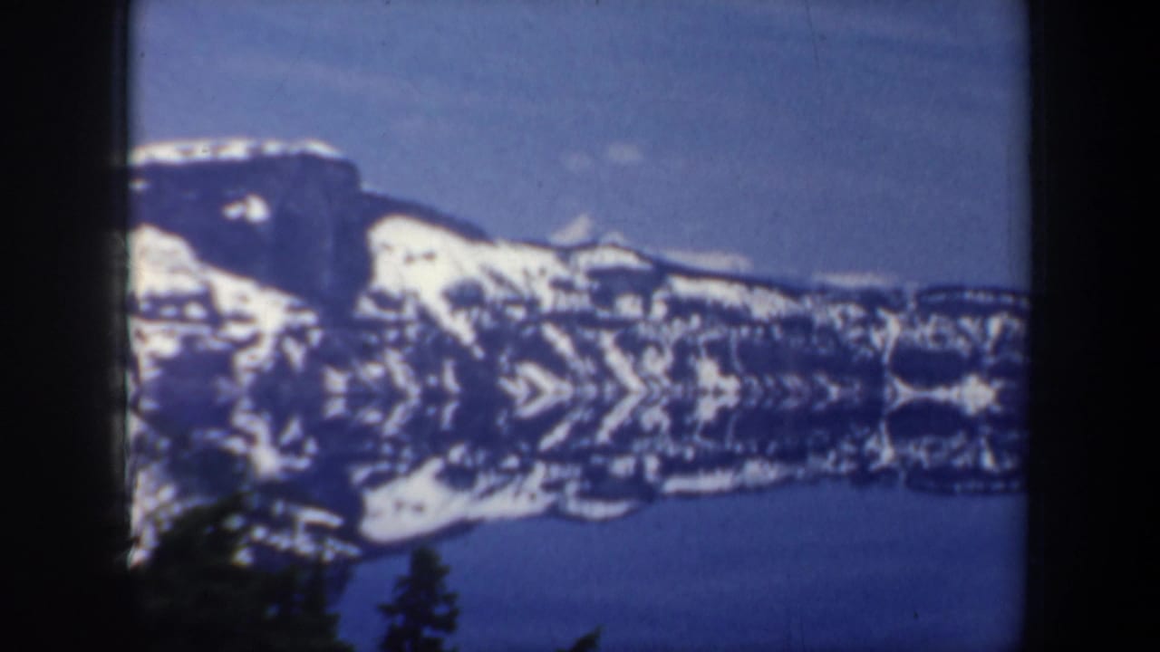Snowy mountain ridge above a calm lake with dark trees in the foreground.