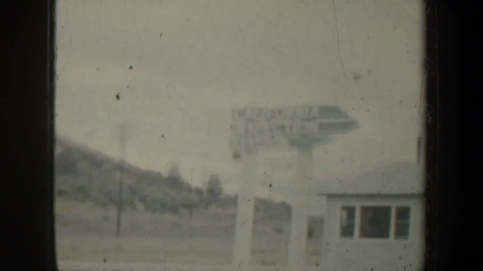 Faded 8mm frame showing a distant California Inspection Station sign beside a small roadside building and hillside.
