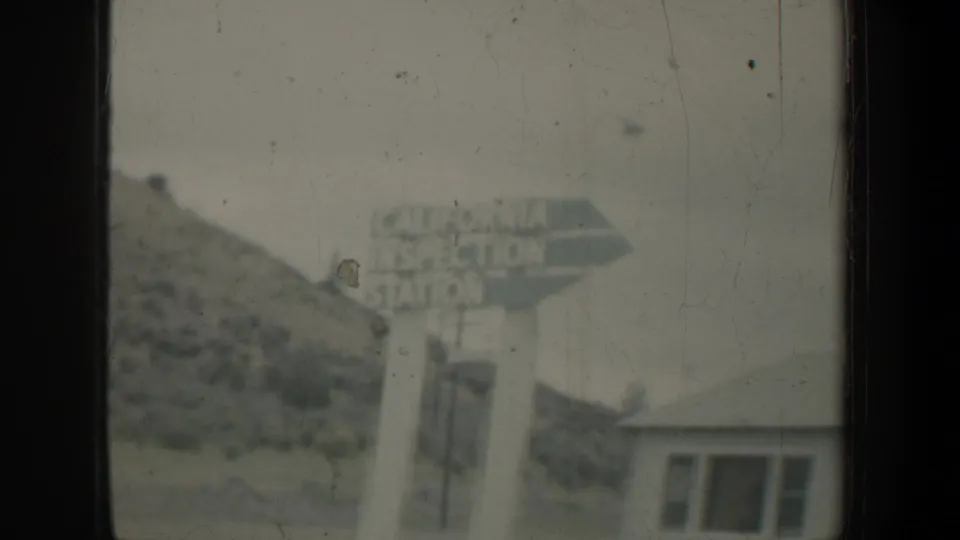 Distant view of a California Inspection Station sign with a small building and hillside in an aged 8mm frame.
