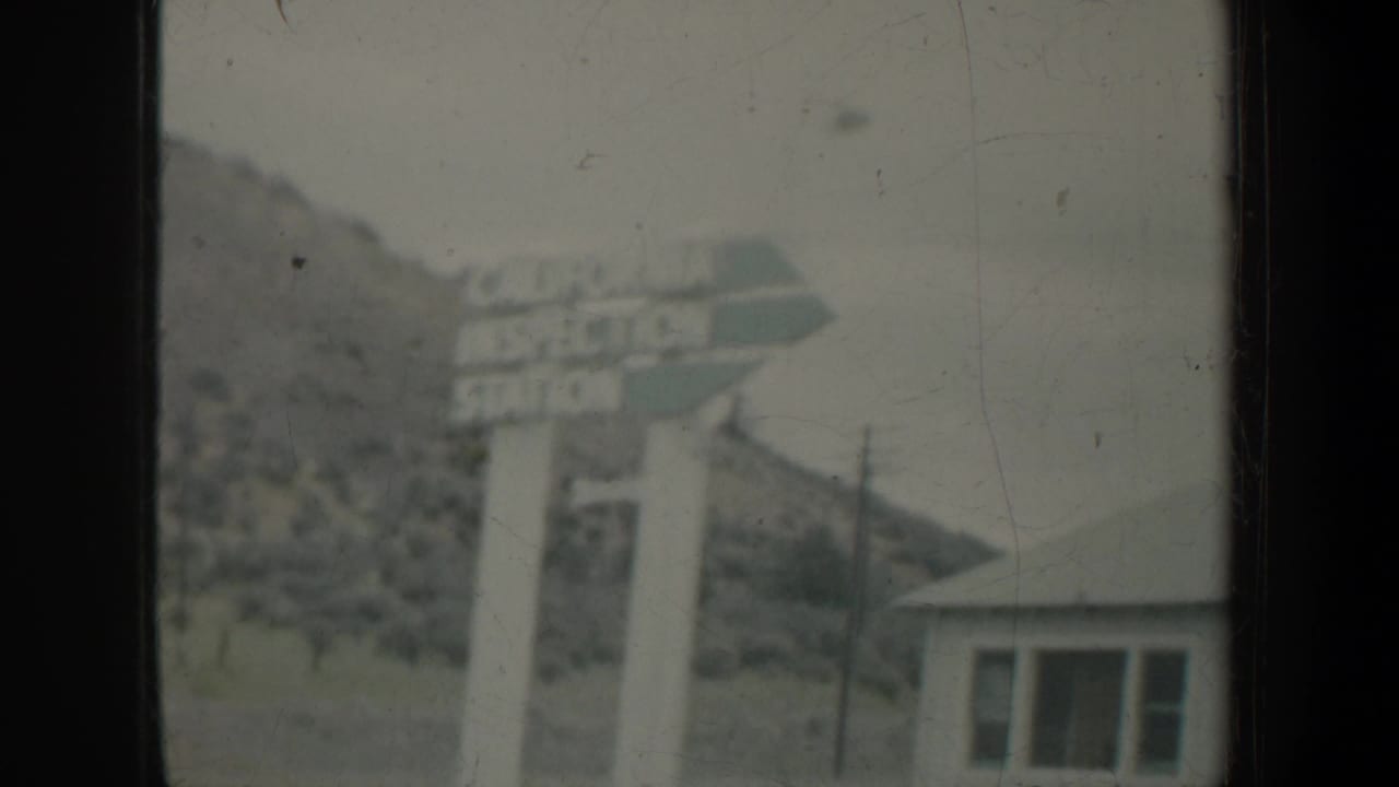 1957 Portland-area home-movie frame showing a roadside sign, small building, hill, and window framing from a moving vehicle.