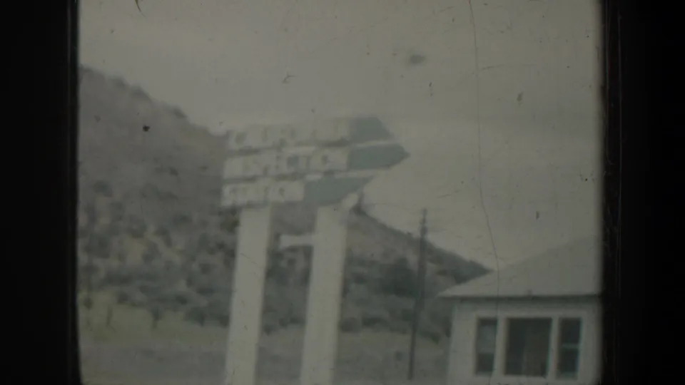 1957 Portland-area home-movie frame showing a roadside sign, small building, hill, and window framing from a moving vehicle.