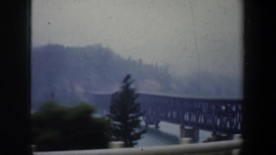 1957 Portland, Oregon frame of a dark truss railroad bridge over a river, viewed through a window with trees and hills beyond.