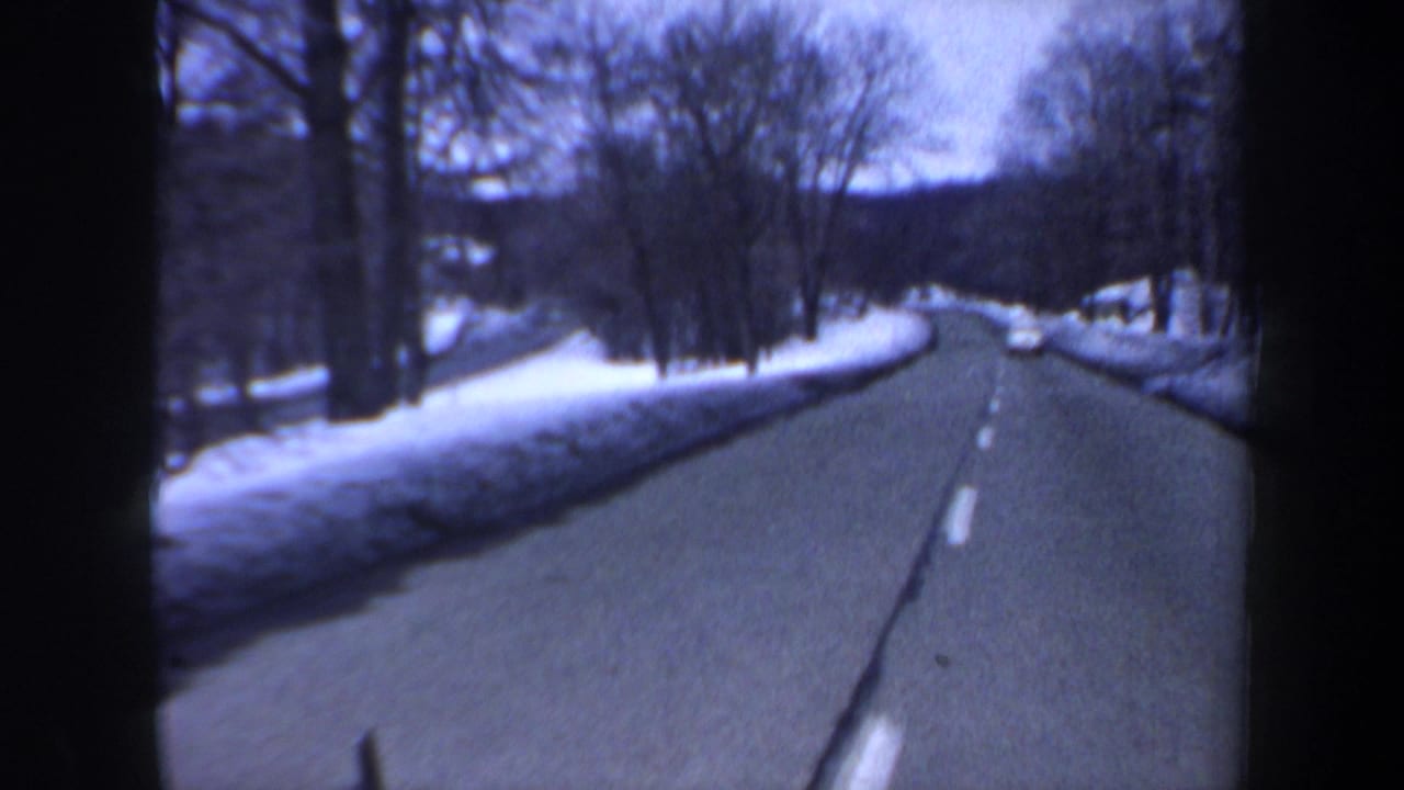 Curving winter road with snowbanks, bare trees, and a vehicle far ahead.