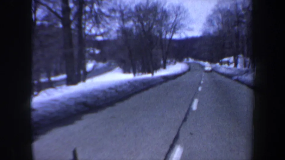A snowy New Hampshire road curves through bare trees in this 1962 home-movie driving view, with a car visible in the distance.