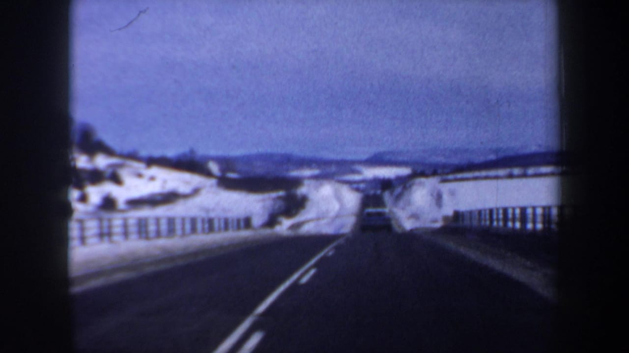 Two-lane snowy highway seen from inside a moving vehicle with a car ahead.