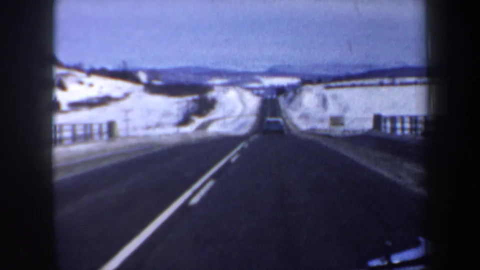 Forward-facing 8mm road view in snowy New Hampshire, showing a dark highway, white center line, guardrails, and one car ahead.