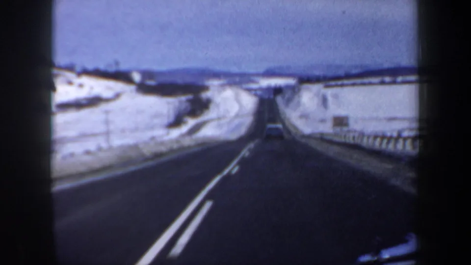1962 8mm home-movie frame from a moving car on a snowy New Hampshire two-lane highway with low hills beyond.