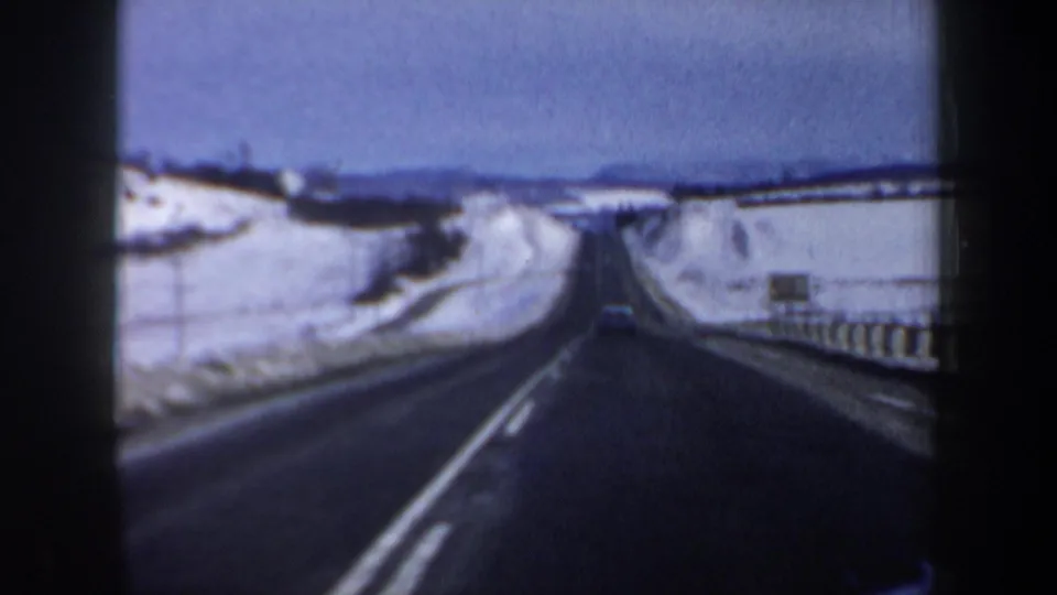 End frame of a 1962 New Hampshire highway drive, with snowbanks, guardrails, lane markings, and a car ahead in soft 8mm color.