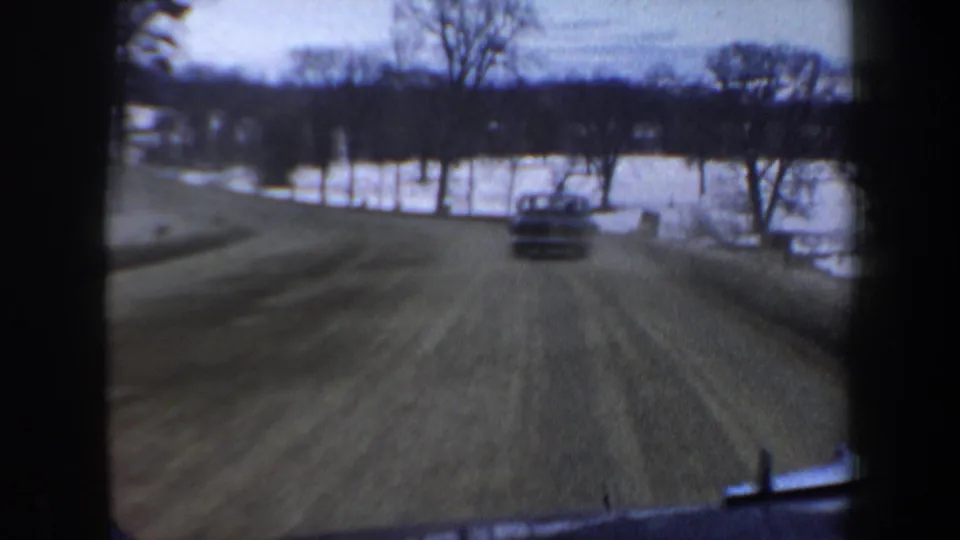 1962 New Hampshire 8mm frame showing a car ahead on a winter rural road, with snow-covered fields and bare trees beyond.