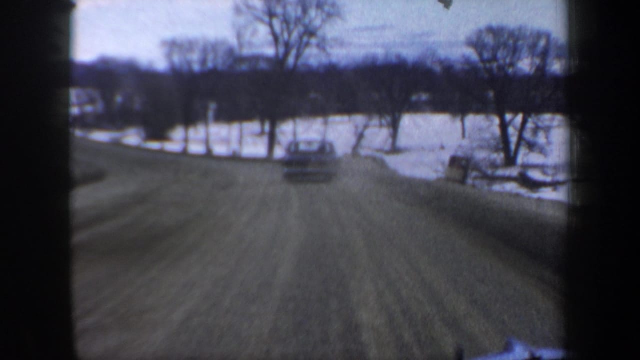 Forward moving-vehicle view in New Hampshire, 1962, following a car along a snowy rural road with trees and open fields.