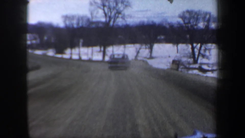 Forward moving-vehicle view in New Hampshire, 1962, following a car along a snowy rural road with trees and open fields.