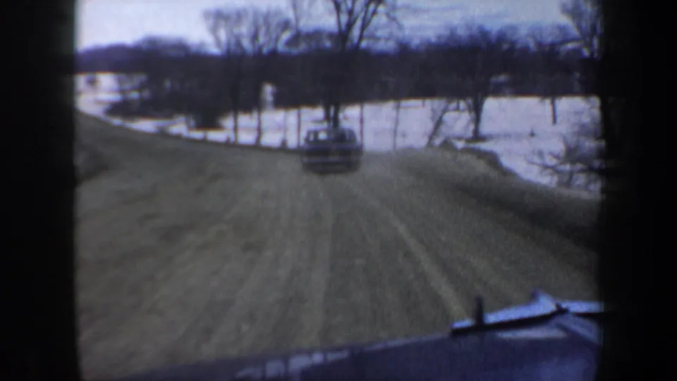 Home-movie frame from inside a moving vehicle, showing a car ahead on a snowy New Hampshire rural road in 1962.