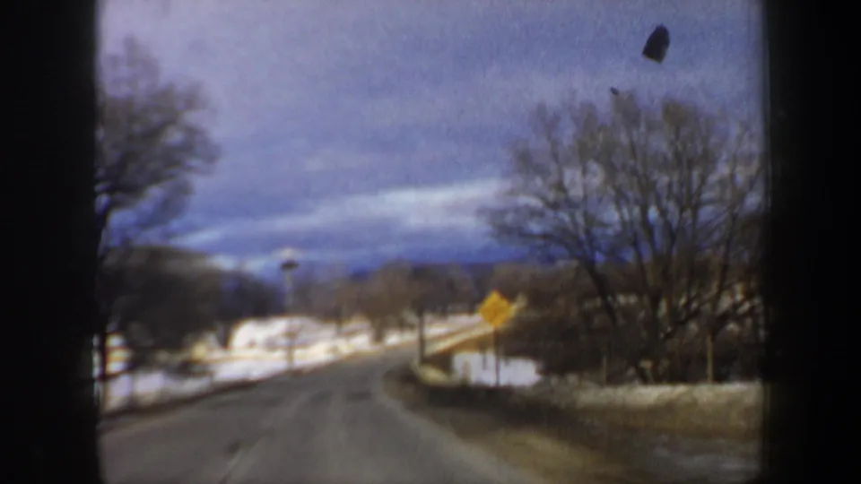 1962 New Hampshire home-movie frame showing a snow-lined rural road curving through bare trees with a yellow roadside warning sign.