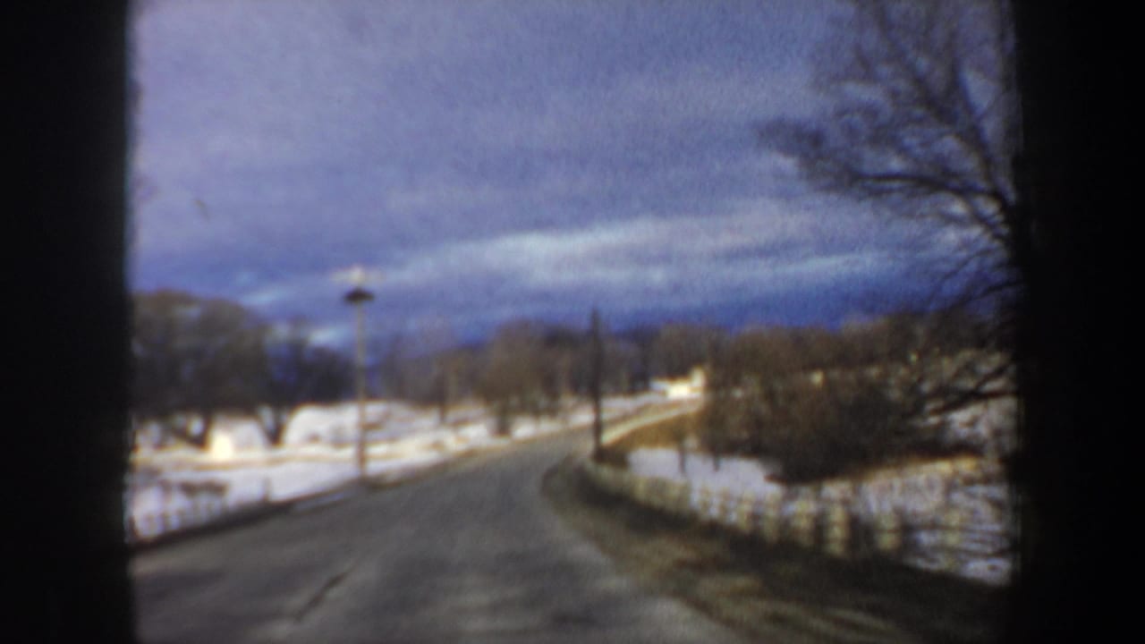 1962 New Hampshire road-view frame with a two-lane snowy roadway, leafless trees, and open winter landscape ahead.
