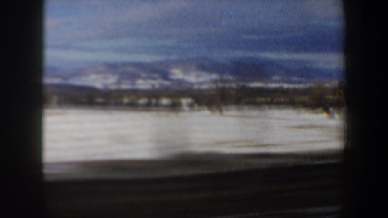 Snow-covered open fields with distant hills seen from a moving vehicle.