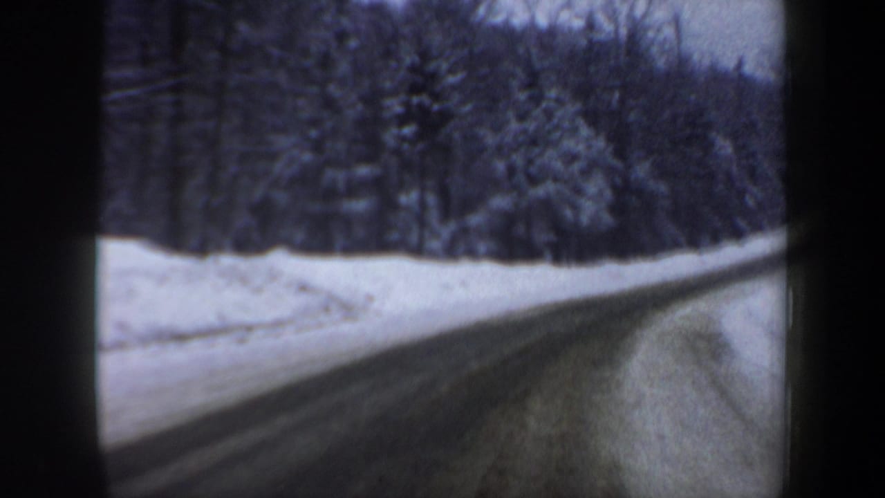 Vehicle-view 8mm frame of an icy New Hampshire highway bordered by snow-covered trees in winter.