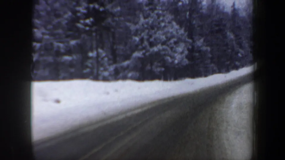 1962 home-movie frame showing a wet or icy rural New Hampshire road running past snowbanks and dense trees.