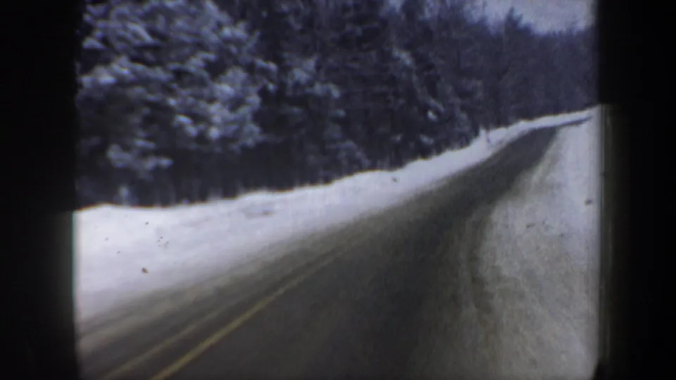 8mm vehicle-view frame of a snow-lined New Hampshire forest highway, with icy pavement and no visible traffic.