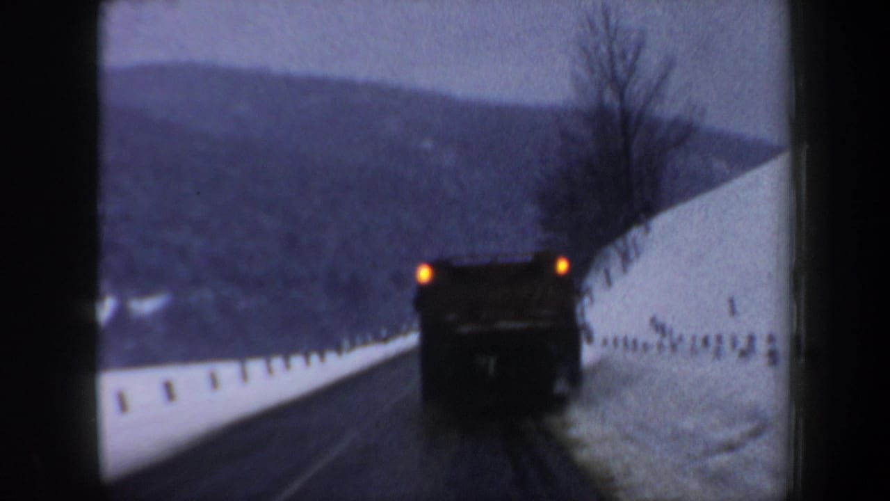 1962 New Hampshire 8mm frame showing a large truck ahead on a curving winter road bordered by snowbanks and hills.