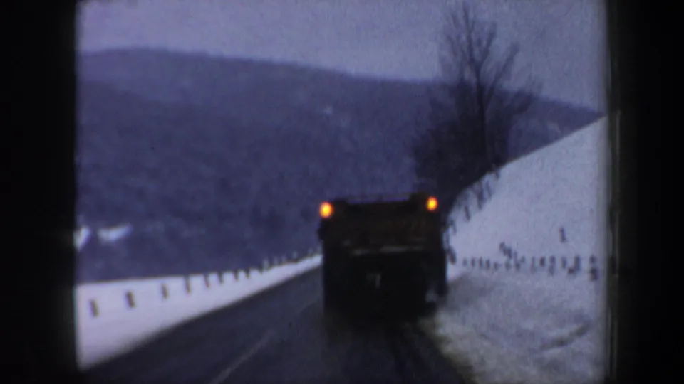 1962 New Hampshire 8mm frame showing a large truck ahead on a curving winter road bordered by snowbanks and hills.