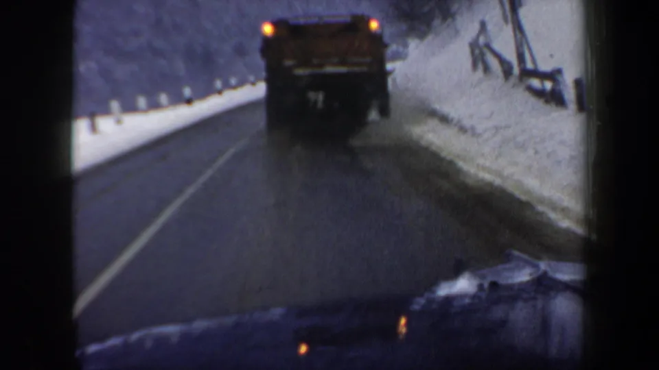 1962 New Hampshire 8mm frame of a road-clearing truck ahead on a wet, winding road with snowbanks and roadside posts.