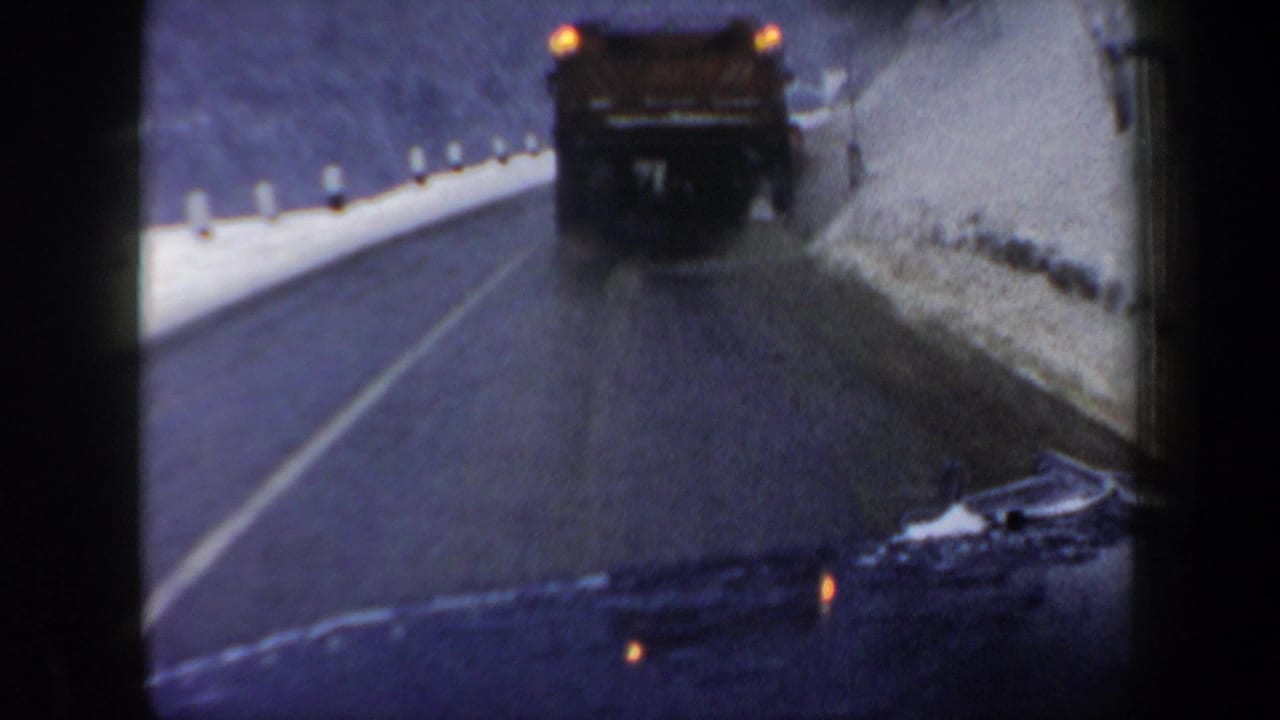 1962 New Hampshire 8mm view from inside a moving car, following a dark truck along a wet road bordered by snowbanks and trees.
