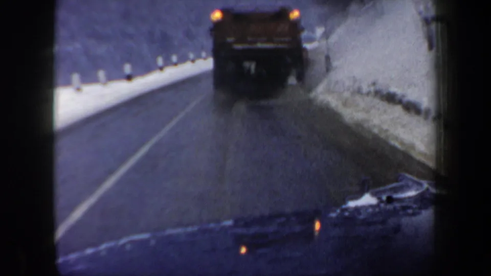 1962 New Hampshire 8mm view from inside a moving car, following a dark truck along a wet road bordered by snowbanks and trees.