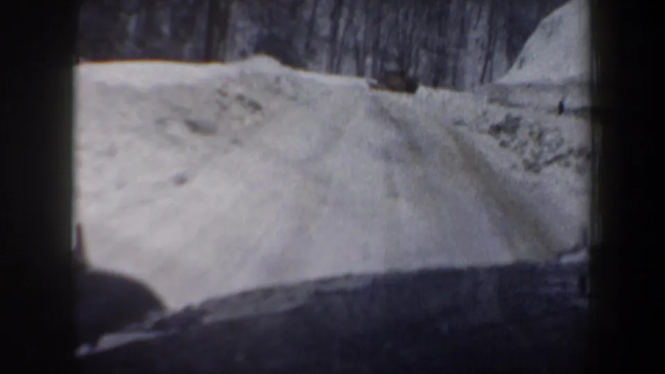 1962 New Hampshire home-movie view from inside a car looking along a narrow snow-covered road with trees and snowbanks ahead.