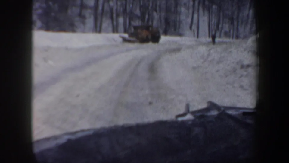 1962 New Hampshire 8mm frame with a plow or truck ahead on a narrow road bordered by deep snow and woods.