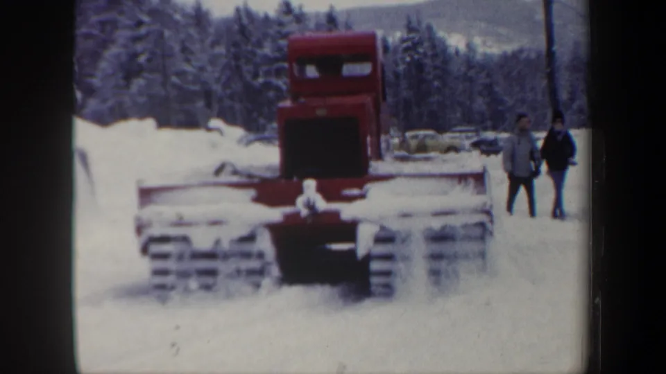 1962 New Hampshire home-movie frame of a red tracked plow in snow, with two bundled observers beside it.