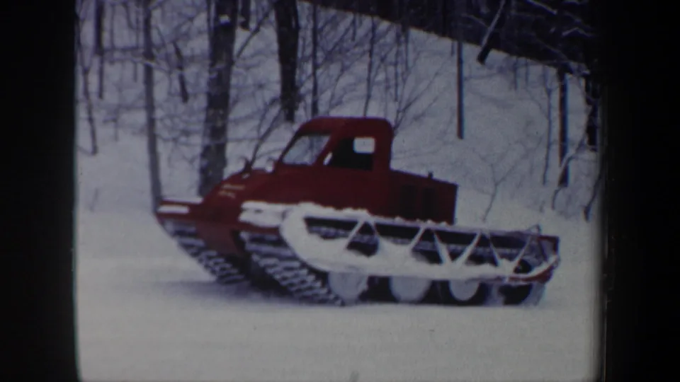 Side view of the red tracked snowplow in a wooded New Hampshire winter scene, with blade and tracks visible in snow.