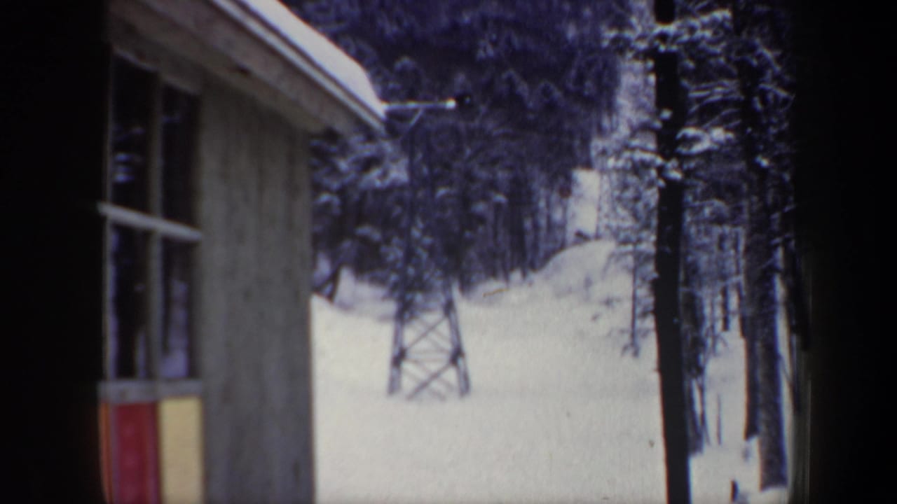 Snowy ski slope seen past a wooden structure, with trees and a metal lift tower outside.