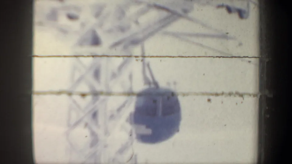 1962 New Hampshire 8mm frame with a blue gondola cable car moving under overhead cables against a pale snowy background.